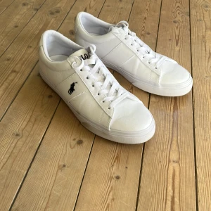 Vita sneakers från Polo Ralph Lauren - Fräscha vita sneakers från Polo Ralph Lauren med klassisk låg profil och rund tå. Skorna har snörning, svart Polo-logga på sidan och texten POLO på hälen. Materialet är en mix av skinn och textil, vilket ger en clean och stilren vibe. Nästintill nya, använda en gång. Nypris 1200 