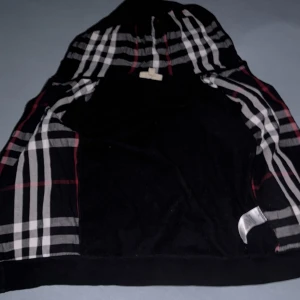Svart Burberry hoodie med rutig huva - Svart hoodie från Burberry med klassisk rutig huva i svart, vitt och rött. Jackan har dragkedja framtill, två fickor och ribbade muddar. Perfekt för dig som vill ha en stilren streetlook med lyxig touch.