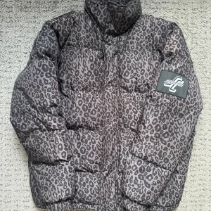 Leopardmönstrad pufferjacka Santa Cruz - Grym pufferjacka från Santa Cruz i mörkgrått leopardmönster. Jackan har hög krage, dragkedja, snörning i nederkant och elastiska muddar vid ärmslut. Perfekt statement-plagg för dig som vill sticka ut. Fodrad insida för extra värme. Skicket är nära till nytt. OBS skickas ej, endast avhämtning i Göteborgsområdet, kan tänka mig att byta mot något Stone Island eller C.P. Company