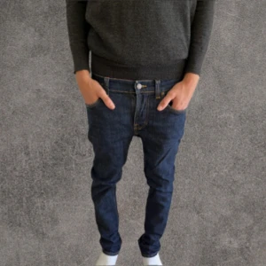 Mörkblå slim jeans från Nudie Jeans W31 L32 - Säljer ett par mörkblå jeans från Nudie Jeans med klassiska orange sömmar och snyggt bakfickemärke. Modellen har normal passform och raka ben, tillverkade i bomull. Perfekta för dig som gillar stilrena och tidlösa jeans.