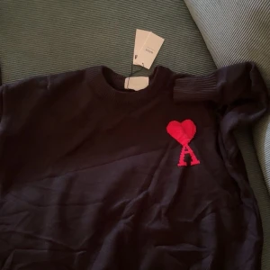 Svart stickad tröja från Ami - Svart stickad tröja från Ami Alexandre Mattiussi med röd hjärtapplikation och bokstaven A på bröstet. Rund hals med ribbade muddar och klassisk passform. Perfekt för dig som gillar stilrena detaljer och vill ha något som sticker ut.