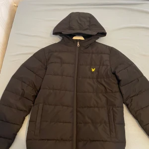 Svart Lyle & Scott pufferjacka - Svart pufferjacka från Lyle & Scott med huva och gul logga på bröstet. Jackan har quiltad design, dragkedja framtill och två sidofickor. Tillverkad i polyester, perfekt för kalla dagar. Enkel och stilren look som funkar till det mesta.