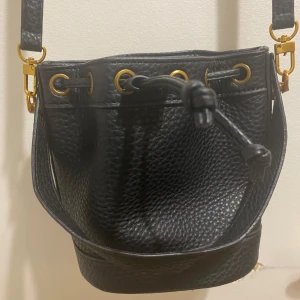 Svart bucket axelväska i skinn (liten modell)  - Snygg svart bucket axelväska i skinn med justerbar rem och guldfärgade metalldetaljer. Väskan har snörning upptill och ett ljust innerfoder med dragkedjefack. Perfekt storlek för att få plats med allt du behöver och cool struktur på ytan. ALDRIG ANVÄND. 