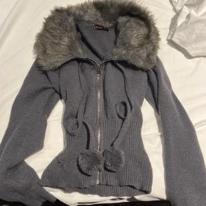 Grå stickad hoodie med pälsluva  - Mysig grå stickad hoodie med fluffig fuskpäls runt huvan. Tröjan har dragkedja framtill, långa ärmar och snörning med två fluffiga bollar. Skriv gärna innan du köper!!