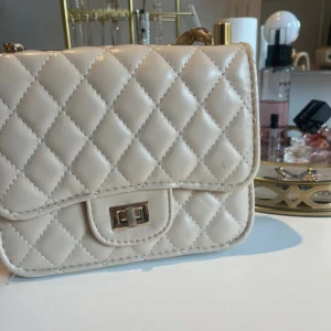 Beige quiltad axelväska med kedja från Ur och Pen - Beige axelväska med quiltat mönster och guldfärgad kedja med insydda detaljer. Väskan har ett vridlås och ett rymligt fack med innerficka med dragkedja. Bra storlek, man får plats med mycket! 