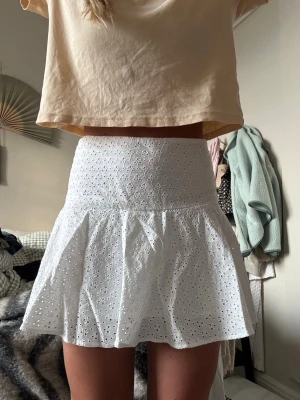 Vit broderad minikjol med volang - Säljer en söt vit minikjol med broderade blommor och volangkant. Kjolen har hög midja och är gjord i ett lätt bomullstyg med hålbroderi som ger en romantisk känsla. Perfekt till sommaren och enkel att matcha med olika toppar.