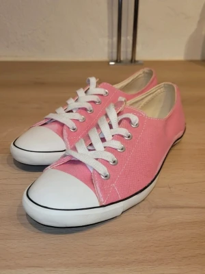 Rosa låga sneakers med vita detaljer - Säljer ett par klassiska låga sneakers i rosa canvas med vita gummisulor och tåhättor. Skorna har vita snören och silverfärgade öljetter. Perfekta för en avslappnad och trendig look.