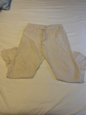 Beiga linnebyxor från H&M - Säljer ett par beiga linnebyxor från H&M med avslappnad passform och snörning i midjan. Perfekta för varma dagar och riktigt sköna att bära. Byxorna har resår i midjan och är gjorda i 100% linne för en luftig känsla. Enda nackdelen är en liten fläck på höger lår 