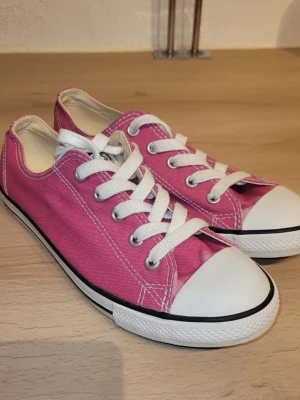 Rosa sneakers med vita detaljer - Säljer ett par klassiska rosa sneakers med vita snören och tåhätta. Skorna har låg profil, vit sula med svart rand och är tillverkade i textilmaterial. Perfekta för dig som vill ha en färgklick till vardagslooken.