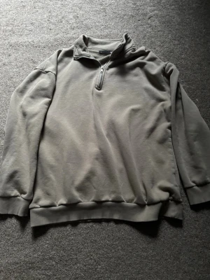 Grå halvzip hoodie - En stilren grå hoodie med halv dragkedja och hög krage. Tillverkad i mjukt sweatshirtmaterial och har ribbade muddar vid ärmslut och nederkant. Perfekt för en avslappnad och enkel look.