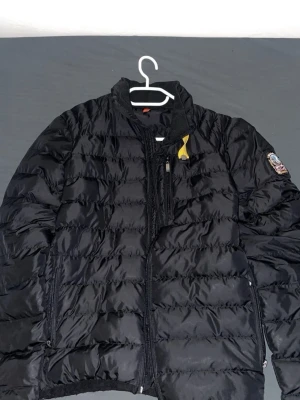 Svart pufferjacka med patch - Säljer en svart pufferjacka med quiltad design och dragkedja framtill. Jackan har en patch på ena ärmen och en gul detalj vid kragen. Perfekt för kalla vinterdagar och har praktiska fickor med dragkedja.Har några små hål