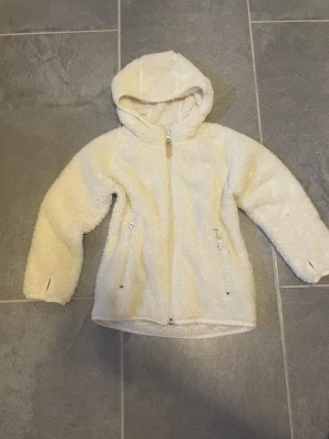 Vit teddyjacka med huva - Mysig vit teddyjacka med dragkedja framtill och två fickor med dragkedja. Jackan har huva och tumhål vid ärmsluten för extra komfort. Perfekt för kyliga dagar när du vill vara både varm och stilren.