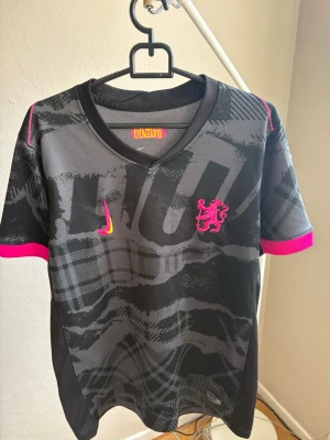 Svart sporttröja med rosa detaljer - Säljer en svart sporttröja med grått mönster och starka rosa detaljer. Tröjan har en rosa lejonlogga på bröstet och rosa Nike-logga. Ärmarna har rosa kant och tyget är lätt och andas, perfekt för träning eller match.