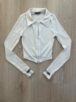 Vit croppad ziptop med textdetalj - Snygg vit croppad topp med dragkedja framtill och krage. Långärmad modell med texten 'PRETTY GIRL' broderad i svart på ärmsluten. Perfekt till jeans eller kjol för en trendig look.