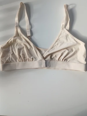 Beige mjuk bh utan bygel - En enkel och bekväm beige bh utan bygel med justerbara axelband och bred resår under bysten. Bh:n har en mjuk trikåkänsla och stängs framtill med hake. Perfekt för dig som gillar en naturlig look och lätt stöd.