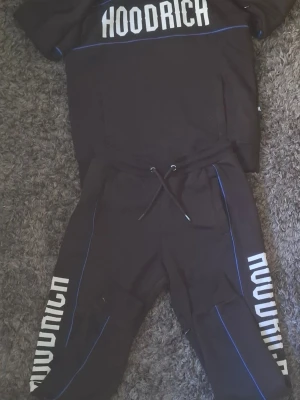 Svart Hoodrich set med blå detaljer - Säljer ett svart Hoodrich set med matchande tröja och byxor. Setet har vita Hoodrich-loggor på både tröja och byxor samt tunna blå linjer som detaljer. Tröjan har huva och korta ärmar, byxorna har dragsko i midjan. Perfekt för en avslappnad streetwear-look.