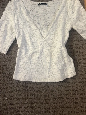 Vit spetsblus från Zara - Säljer en superfin vit blus från Zara med v-ringning och trekvartsärm. Blusen är täckt av spets med blommigt mönster och har en feminin, lätt figurnära passform. Perfekt att styla med jeans eller kjol för en trendig look.