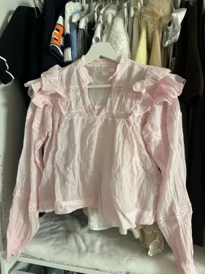 Ljusrosa volangblus från Gina Tricot - Supersöt ljusrosa blus från Gina Tricot med volanger på axlarna, spetsdetaljer och v-ringning. Blusen har långa ärmar och är gjord i ett tunt, krinklat tyg som ger en avslappnad men romantisk look. Perfekt för dig som gillar en feminin stil.