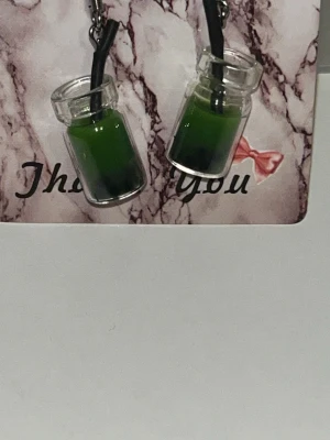Örhängen med gröna bubble tea-flaskor - Unika örhängen formade som små glasflaskor fyllda med grön vätska och svarta pärlor, inspirerade av bubble tea. Flaskorna har ett svart sugrör och hängs i metallfästen. Perfekt accessoar för dig som gillar roliga och kreativa smycken. Örhängens krokarna är i rostfritt stål 