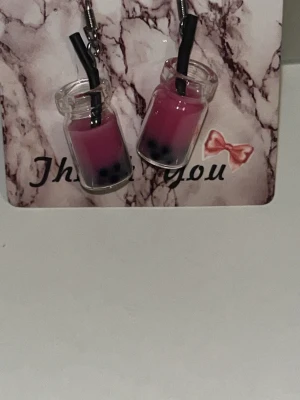 Bubble tea örhängen i rosa  - Unika örhängen formade som små bubble tea-glas med rosa  dryck, svarta sugrör och svarta pärlor som ser ut som tapiokabollar. Tillverkade i glas och metall, perfekta för dig som älskar roliga accessoarer. Örhängens krokarna är i rostfritt stål 