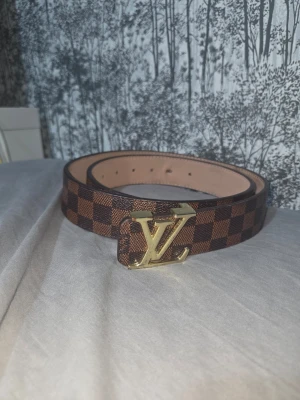 Brunt rutigt LV-bälte med gulddetalj - Snyggt bälte i brunt med rutigt mönster och stor guldfärgad LV-logga som spänne. Bältet har en klassisk design och är gjort i ett läderliknande material med beige insida. Perfekt för att lyfta vilken outfit som helst.