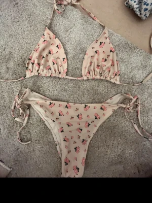 Beige bikini med blommigt mönster - Supersöt beige bikini med små rosa blommor och gröna blad. Överdelen har trekantskupor med knyt i nacken och ryggen, och underdelen har knyt i sidorna för perfekt passform. Perfekt för lata dagar på stranden eller poolen!