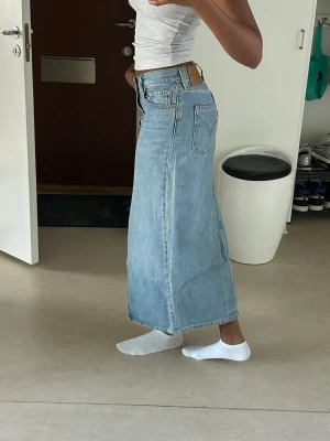 Levis kjol - Säljer en ljusblå lång jeanskjol i klassisk denim med hög midja och rak passform. Kjolen har en snygg slits framtill och femficksmodell. 