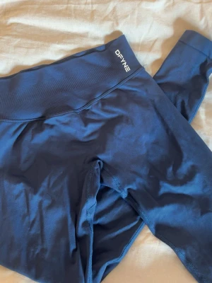 Mörkblå seamless leggings från DFYNE - Mörkblå impact leggings från DFYNE med låg/medel midja och bred resår. Tillverkade i 90% nylon och 10% elastan för en stretchig och bekväm passform. Snygg logga på linningen och texten 'LET NOBODY DFYNE YOU' på insidan. Använda 2gånger, säljer för dem inte kommer till användning! Nypris: 673kr