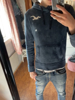 Hollister hoodie - Snygg blå hoodie från Hollister med vit logga på bröstet och stort HOLLISTER-tryck längs ena ärmen. Hoodien har huva med dragsko och en stor magficka. Perfekt för en avslappnad och trendig stil.