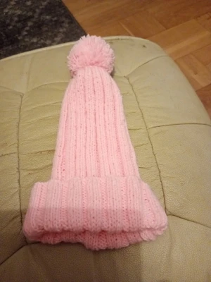 Ljusrosa stickad mössa med tofs - Säljer en söt ljusrosa stickad mössa med bred uppvikt kant och en fluffig tofs på toppen. Mössan har ribbstickat mönster och är gjord i mjukt garn som håller dig varm under kalla dagar. Perfekt accessoar för att addera lite färg till din vinterstil. 100% Polyester. Tvättråd 40 grader. Hemstickad.