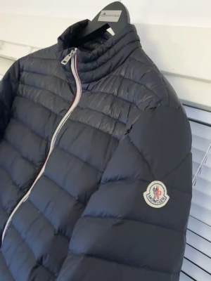 Svart dunjacka från Moncler storlek 3 - Snygg svart dunjacka från Moncler med quiltad design och raka ärmar. Jackan har dragkedja framtill med Monclers signaturfärger och en matchande dragkedja på fickan. Märkeslogga på vänster ärm.