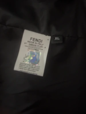 Svart jacka från Fendi i storlek XL - Snygg svart jacka från Fendi i storlek XL. Jackan är tillverkad i Italien och har en klassisk design med Fendi-logga på insidan. Materialet känns som polyester och jackan har en mörk ton som passar till många outfits.