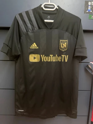 Svart LAFC matchtröja från Adidas - Snygg svart LAFC matchtröja från Adidas med guldiga detaljer och YouTube TV-logga på bröstet. Tröjan har klassiska Adidas-ränder på axeln, klubbmärke och är tillverkad i lätt funktionsmaterial (Aeroready). Perfekt för fotboll eller träning.