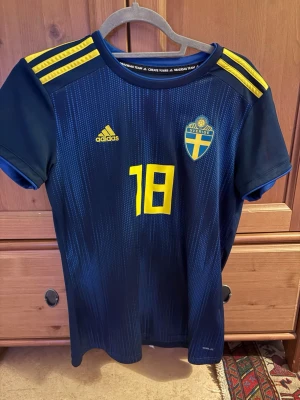 Blå Sverige fotbollströja Adidas - Säljer en blå fotbollströja från Adidas med Sveriges landslagsemblem och nummer 18 i gult. Tröjan har gula detaljer på axlarna och korta ärmar. Perfekt för match eller träning. Tillverkad i lätt och ventilerande material.