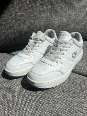 Vita låga sneakers från Champion - Fräscha vita sneakers från Champion med klassisk låg profil och grå detaljer. Skorna har snörning, diskret logga på sidan och ovandel i syntetmaterial och mesh för extra komfort. Perfekta för en clean och sportig look.