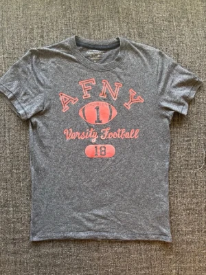 Grå t-shirt med tryck från Abercrombie - Snygg grå t-shirt från Abercrombie & Fitch med rött tryck framtill. Texten 'AFNY Varsity Football' och en amerikansk fotboll ger en sportig vibe. Klassisk rund hals och korta ärmar. Perfekt till jeans eller shorts.
