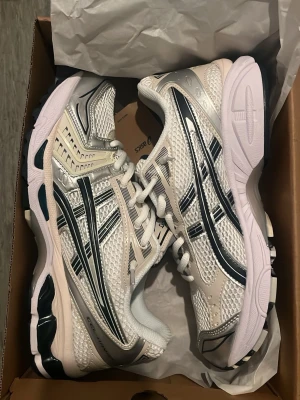 Asics gel kayano 14 Vita och silver  - Snygga sneakers från Asics i vitt och silver med svarta detaljer. Skorna har en sportig mesh-överdel för bra ventilation och klassiska Asics-loggor på sidorna. Sulan är vit med svarta inslag och ger bra stöd. Perfekta för en trendig och bekväm stil.