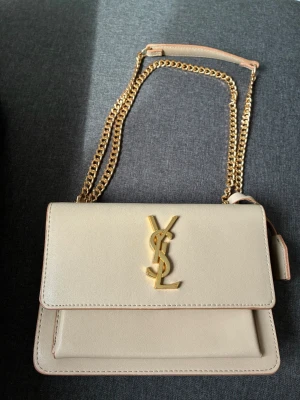Beige Saint Laurent handväska i skinn - Snygg beige handväska från Saint Laurent i slätt skinn med guldfärgad kedjerem och ikonisk YSL-logga framtill. Väskan har ett stilrent och elegant uttryck med praktisk axelrem i metall och läderdetaljer.