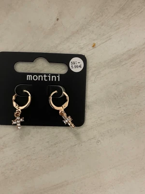 Guldiga örhängen med kors från Montini - Säljer ett par små guldiga hoops från Montini med hängande kors dekorerade med glittrande stenar. Perfekta för dig som gillar detaljer och vill ha något som sticker ut lite extra. Snygg accessoar till alla outfits!