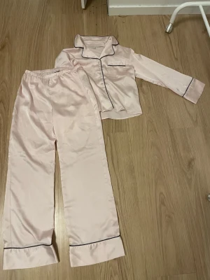 Ljusrosa satinpyjamas med svarta kanter - Snygg tvådelad pyjamas i ljusrosa satin med svarta kontrastsömmar. Setet består av en långärmad skjorta med krage och bröstficka samt matchande byxor med resår i midjan. Perfekt för mysiga kvällar hemma.