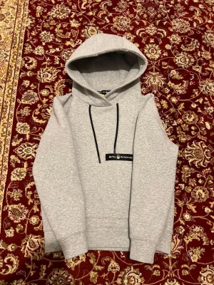 Grå hoodie Sail Racing - Snygg grå hoodie från Sail Racing med svart logga på bröstet och svarta dragsnören i huvan. Klassisk passform med ribbade muddar vid ärmar och nederkant. Perfekt för en chill och avslappnad stil. Storlek XS 