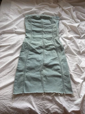 Ljusblå axelbandslös jeansklänning - Säljer en ljusblå axelbandslös jeansklänning med rå kant nertill. Klänningen har en figurnära passform och markerade sömmar som ger en snygg siluett. Perfekt för dig som gillar en edgy och trendig stil. Använd endast en gång. Nypris 500 kr, mitt pris 249 kr. 