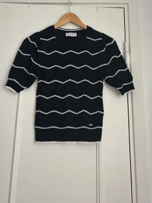Svart stickad topp från Holly & Whyte - Snygg svart stickad topp från Holly & Whyte med vita vågiga ränder och ribbade kanter. Modellen har trekvartsärm och dekorativa bollar längs ärmslut och nederkant. Perfekt till jeans eller kjol för en trendig look.