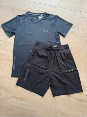Under Armour träningsset svart - Snyggt träningsset från Under Armour med en svart t-shirt och matchande shorts. Tillverkade i ett lätt och snabbtorkande material som passar perfekt för gymmet eller löpning. T-shirten har rund hals och logga på bröstet, shortsen har resårmidja och logga på benet.