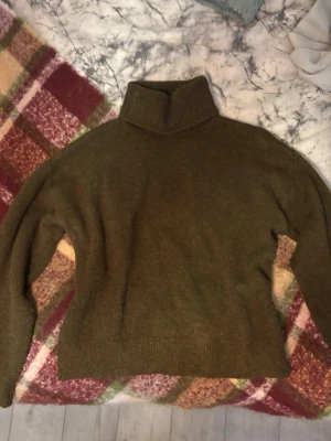 Olivgrön polotröja i ullmix - Mysig olivgrön stickad polotröja med hög krage och avslappnad passform. Tröjan har långa ärmar och ribbade muddar vid ärmslut och nederkant. Perfekt för kalla dagar och enkel att styla till olika outfits.