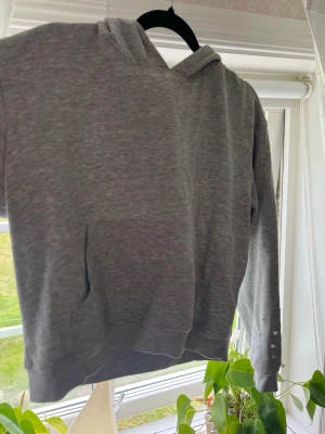 Grå hoodie med pärlor på ärmen - Säljer en grå hoodie med huva och magficka. Ärmen är dekorerad med vita pärlor som ger en unik och trendig look. Perfekt för dig som vill ha något extra till vardagsstilen. Mjuk och skön bomullsblandning.