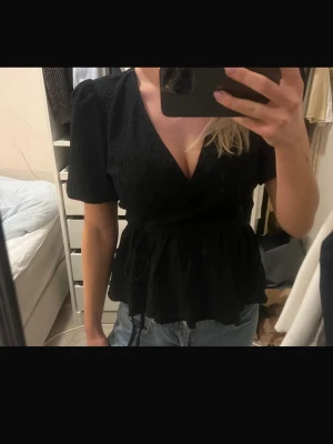 Svart omlottopp med knyt - Säljer en svart omlottopp med knyt i midjan och korta puffärmar. Toppen har v-ringning och en lätt volang nedtill som ger en feminin siluett. Perfekt att styla med jeans eller kjol för en trendig look. Använd fåtal gånger 