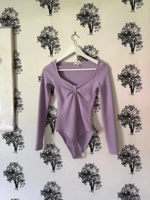 Ljuslila ribbad body med lång ärm - Säljer en ljuslila body med lång ärm i ribbat material. Bodyn har en djup, rundad urringning och en söt knutdetalj framtill. Materialet ser stretchigt och mjukt ut, perfekt för en trendig och bekväm look.