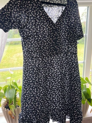 Blå blommig kortklänning - Snygg blå kortklänning med vitt blommönster och v-ringning. Klänningen har korta ärmar och volangdetalj nertill. Perfekt för varma dagar och enkel att styla med sneakers eller sandaler.