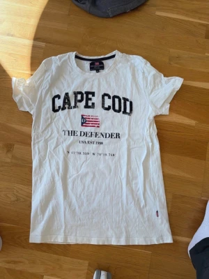 Vit CAPE COD t-shirt med tryck medium  - Säljer en vit t-shirt med svart text 'CAPE COD' och tryck framtill. T-shirten har rund hals och korta ärmar. Materialet känns mjukt och skönt, perfekt till sommaren. Snygg och enkel att matcha med jeans eller shorts. Medium